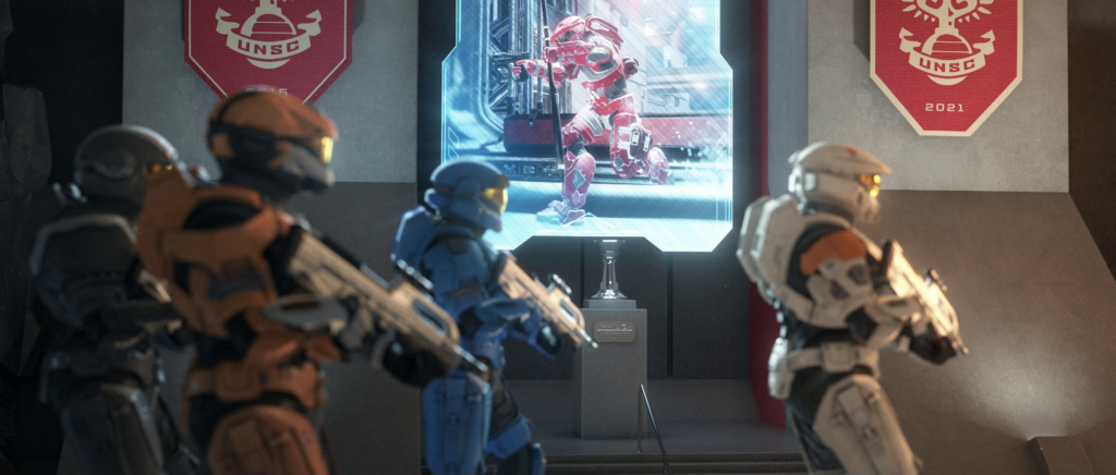 4 Halo Infinite Spartans in the foreground out of focus and in the background is a screen playing a famous moment from a Halo 5 match with the Halo World Championship trophy resting in front of it as a museum exhibit.