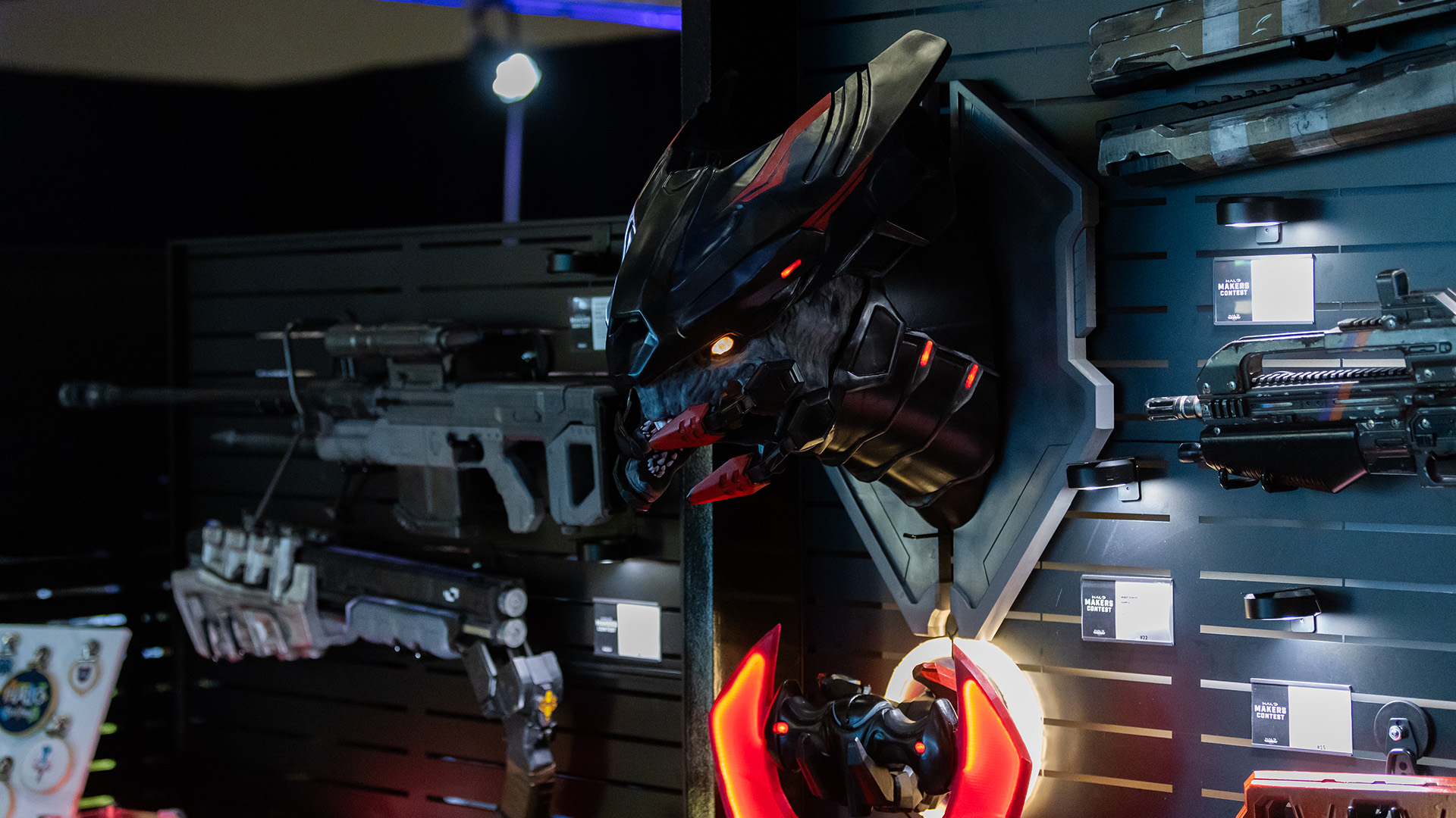 Creations from the Halo World Championship Maker's Contest hang displayed on a wall. A mounted trophy head of Jega 'Rdomnai hangs above a scale replica of an Elite Bloodblade, surrounded by more replica Halo weaponry.